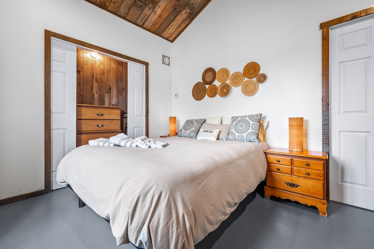 The bedroom features a spacious bed with a light-colored cover and decorative pillows. Warm wooden accents are visible on the walls and ceiling. Two wooden bedside tables with lamps are positioned beside the bed. A dresser with multiple drawers is also in the room.