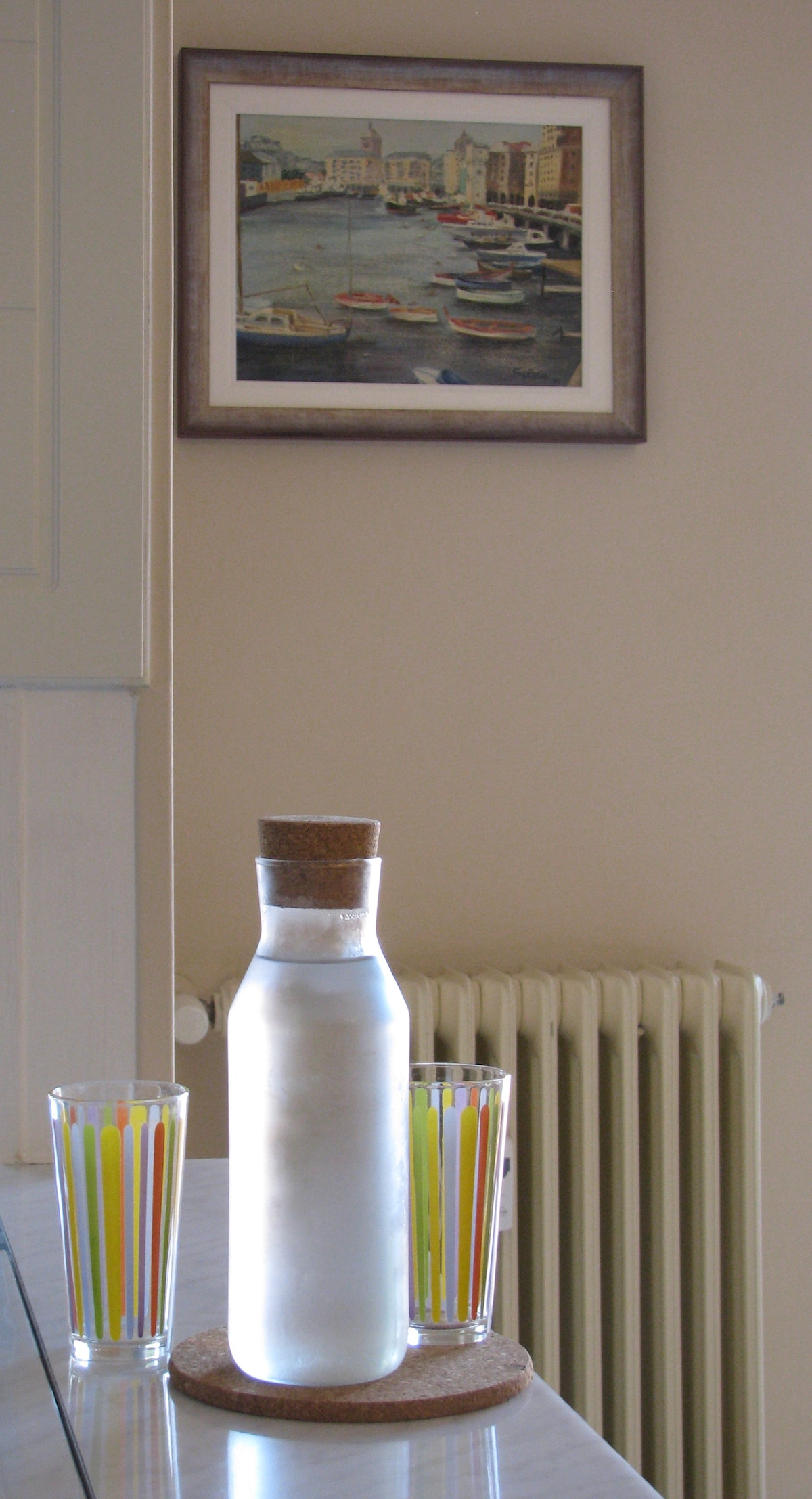 A glass pitcher with a cork lid is placed on a circular cork mat. Two colorful drinking glasses with vertical stripes sit beside it. In the background, a framed artwork depicting a harbor scene with boats is hung on the wall, while a radiator is subtly visible.