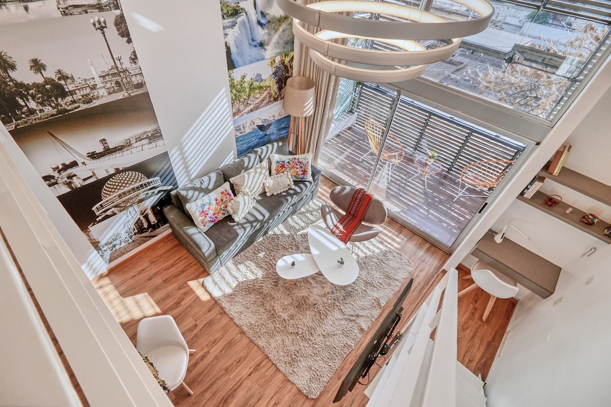 A spacious living area is featured, furnished with a sectional sofa adorned with colorful pillows. A round coffee table and plush area rug create a cozy atmosphere. Bright natural light streams through large windows, complementing the stylish decor with a backdrop of iconic city imagery.