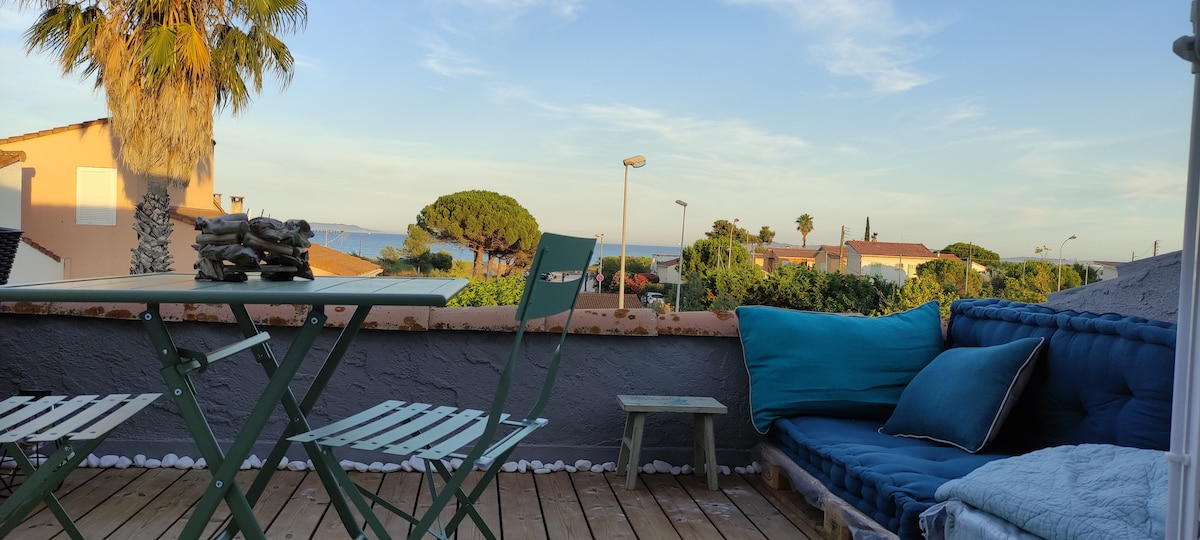 A terrace is showcased with comfortable seating and a small dining area. A blue couch is accompanied by a green table and chairs, all set against a backdrop of greenery and a distant view of the sea under a clear sky.