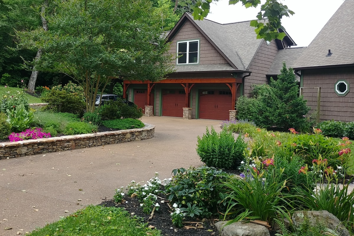 An inviting entrance showcases a well-maintained driveway leading to a rustic guesthouse, framed by lush landscaping. Colorful flowers in various hues enhance the curb appeal, while stone accents add texture to the garden beds, creating a welcoming atmosphere amidst the surrounding greenery.