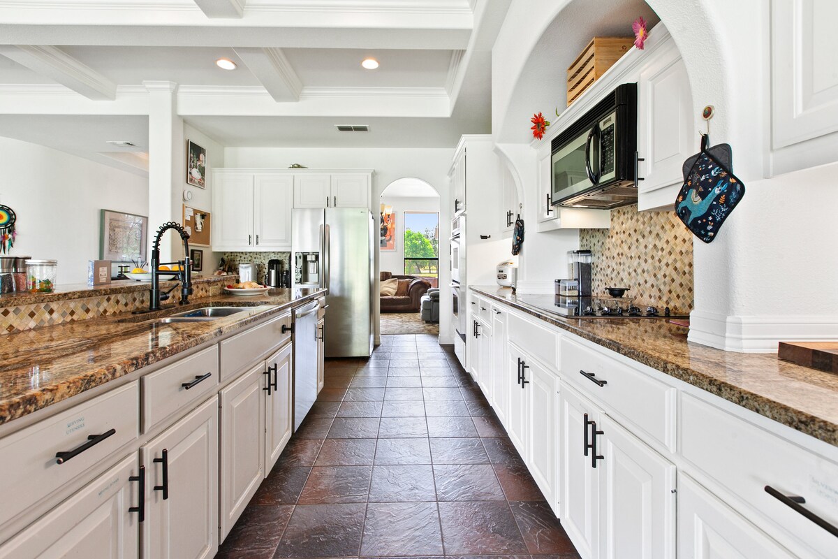 An open concept kitchen is depicted with white cabinetry and granite countertops. Two long countertops line each side, offering ample workspace. Stainless steel appliances provide modern convenience. Natural light floods the space through windows, enhancing the inviting atmosphere of the room.