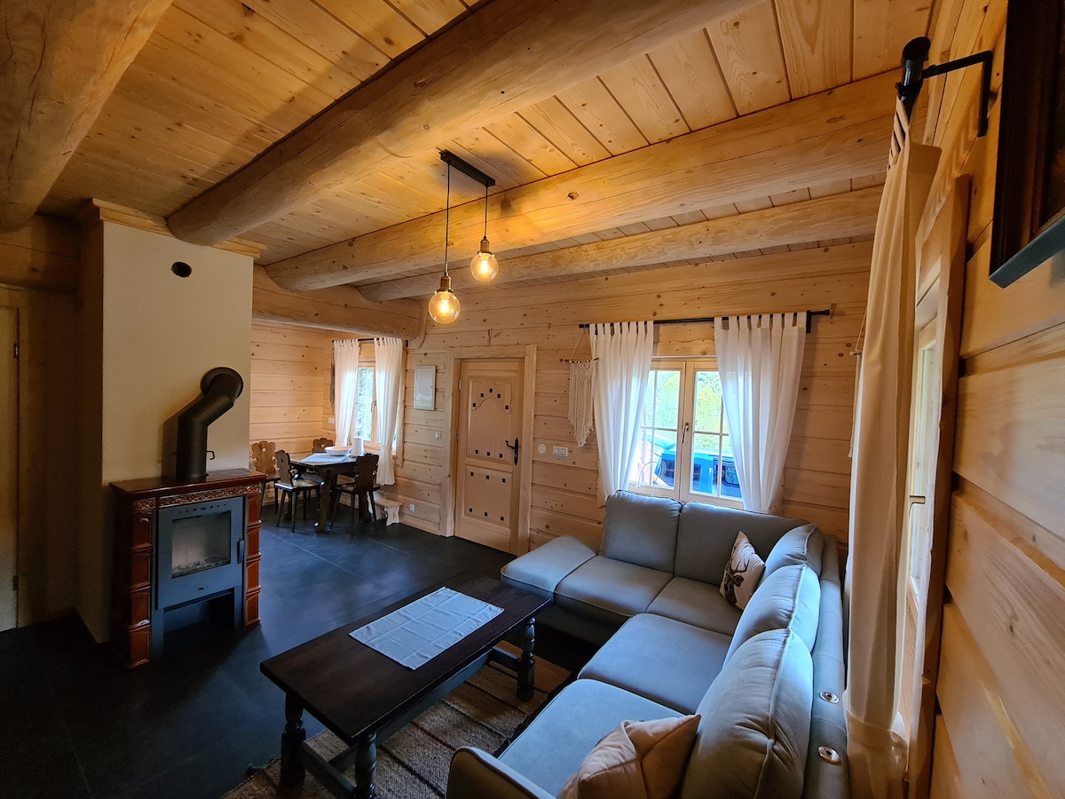 An inviting living space features wooden walls and ceiling beams, combining rustic charm with modern comfort. A sectional sofa faces a low coffee table, while a dining table for four is situated nearby. Soft, sheer curtains frame the windows, and a wood stove adds warmth to the room.