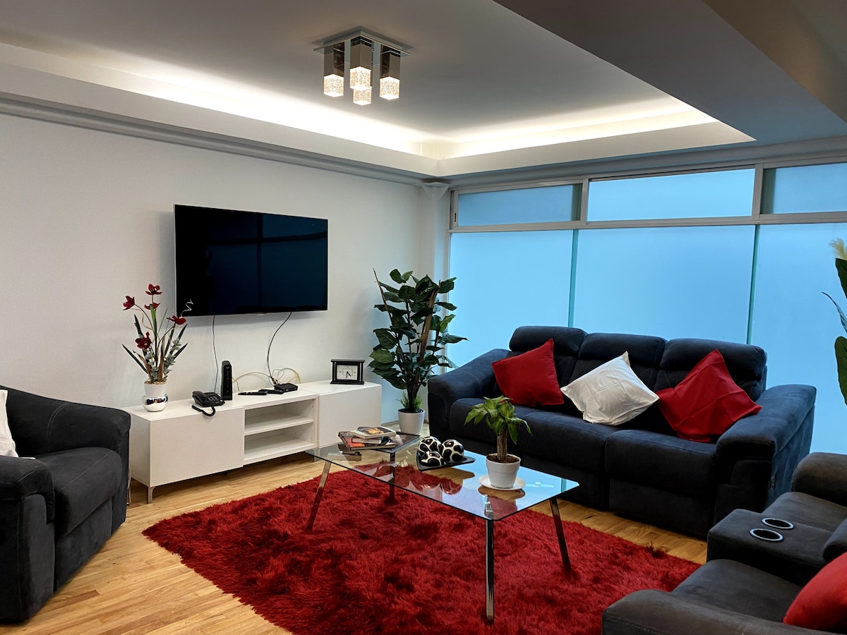 A modern living area features a gray sectional sofa with red accent pillows, complemented by a glass coffee table. A large flat-screen TV is mounted on the wall, and a vibrant red rug enhances the warm ambiance. Natural light streams in through large windows.