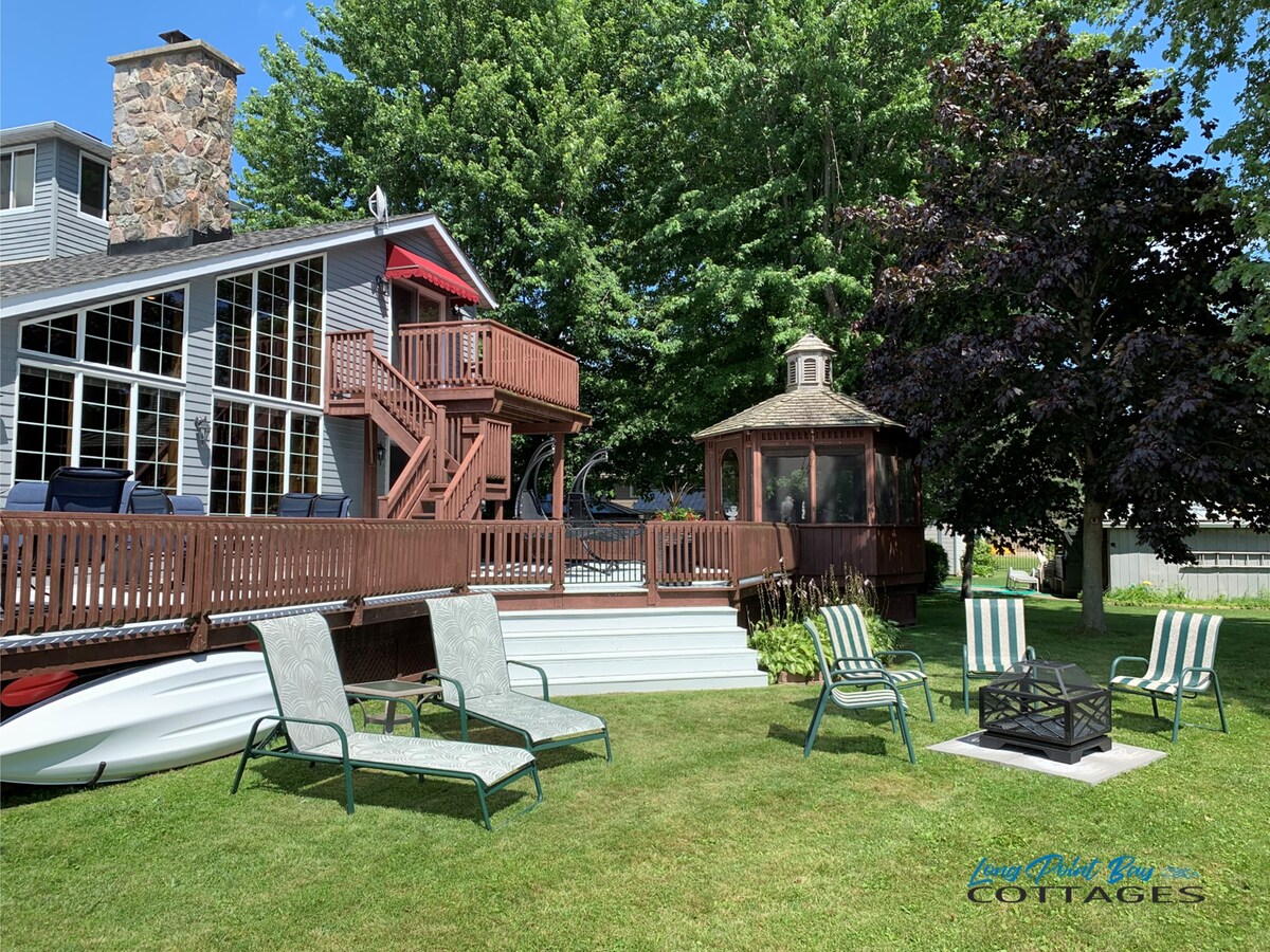 A spacious outdoor area is shown, featuring a large deck attached to the cottage and a gazebo among lush greenery. Several lounge chairs are arranged around a firepit, inviting relaxation and social gatherings. A row of steps leads up to the upper deck, enhancing accessibility.
