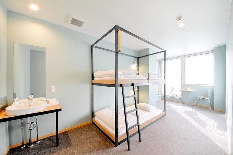 A shared dormitory room features a sturdy bunk bed with crisp white linens. A compact washbasin is positioned nearby. The room is illuminated by natural light from large windows, complemented by a small table and chairs for relaxation or work.
