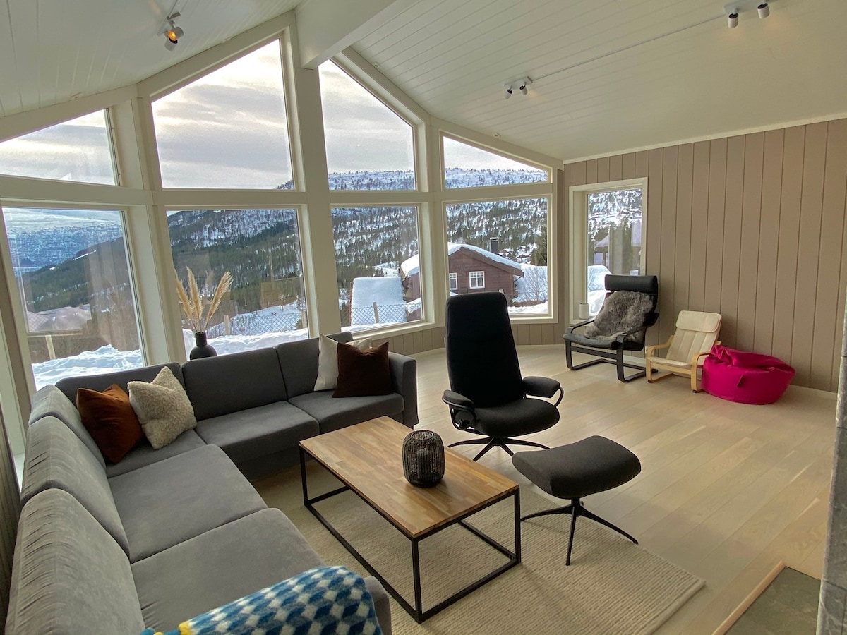 A sunlit living area features large windows providing panoramic mountain views. A spacious sectional sofa and lounge chair are arranged around a wooden coffee table. A soft area rug and decorative items create a cozy environment, while natural light enhances the inviting atmosphere.