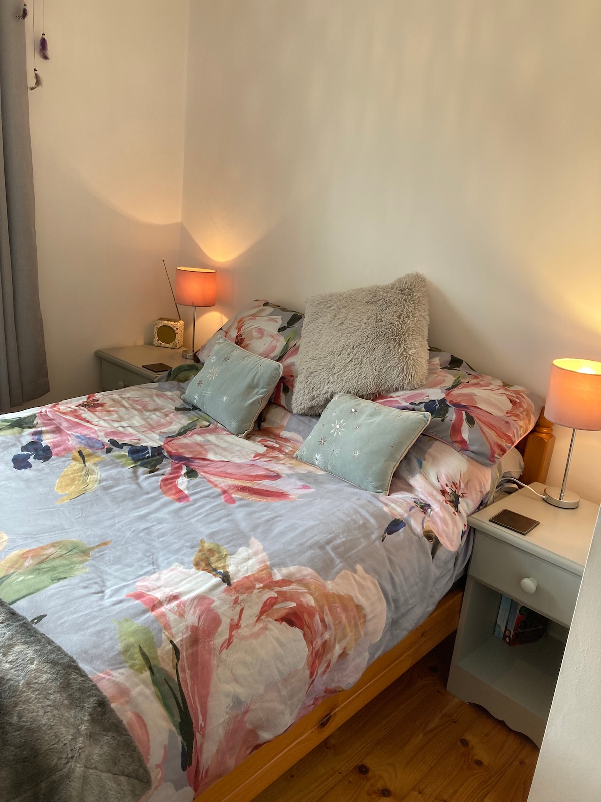 A cozy sleeping area features a double bed adorned with a floral comforter and decorative pillows. Soft lighting is provided by two bedside lamps, creating a warm ambiance. Light-colored walls complement the wooden flooring, enhancing the room's inviting feel.