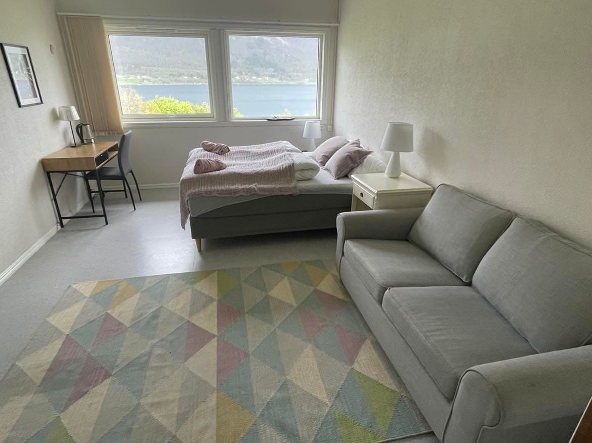 A bright sleeping area features a spacious bed with light bedding, complemented by two bedside lamps. A cozy sofa sits nearby, and a desk with a chair is positioned next to a large window that offers views of the surrounding nature and water.