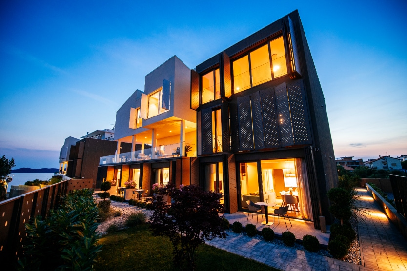 The exterior of the villa is illuminated at dusk, highlighting modern architectural features and expansive glass windows. A landscaped garden with neatly trimmed bushes is positioned in the foreground, enhancing the serene atmosphere of this luxury property by the sea.