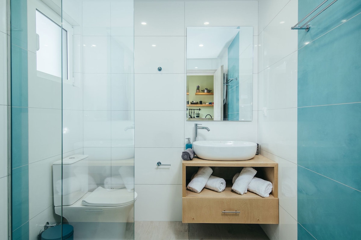 A modern bathroom showcases sleek white tiles and turquoise accents. A spacious vanity features a round sink and a wooden cabinet with neatly folded towels. A glass shower enclosure offers an open and airy feel, with natural light filling the space from a nearby window.
