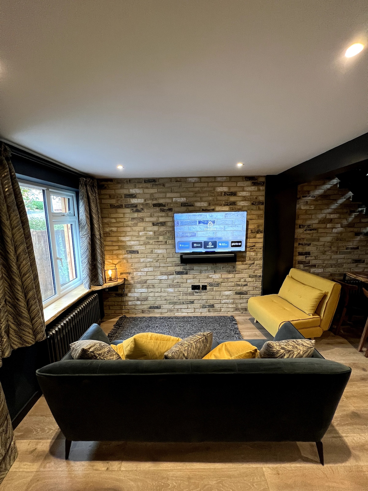 An inviting living area features a dark sofa with patterned throw pillows facing an interactive TV mounted on a textured brick wall. Natural light streams through a large window adorned with elegant curtains, complementing a cozy atmosphere created by warm lighting.