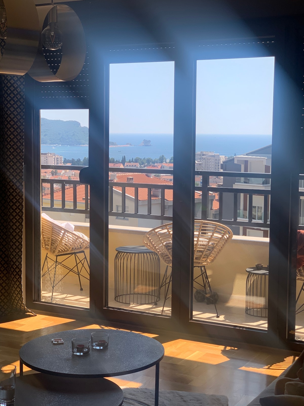 Large glass doors reveal a stunning view of the sea and nearby island. Outside, two modern chairs and a table are positioned on a balcony. Sunlight streams in, enhancing the light wood flooring and the cozy atmosphere of the living space.
