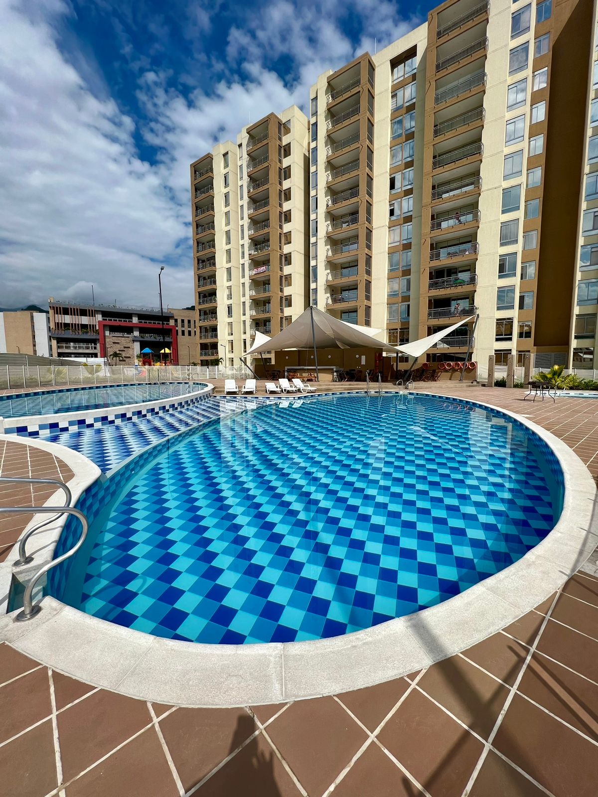A vibrant private pool is surrounded by a tiled area featuring umbrellas and lounge chairs. The pool's blue and white checkered tiles create a striking visual effect. A five-story building is visible in the background, with large glass windows and a sunny sky above.