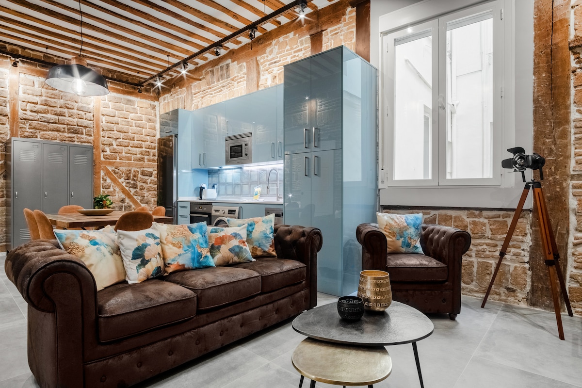 A spacious living area features a brown leather sofa adorned with decorative pillows, positioned across from a stylish kitchen with blue cabinetry. Natural light enters through a large window, illuminating the modern decor, which includes a round coffee table and wooden dining chairs in the background.