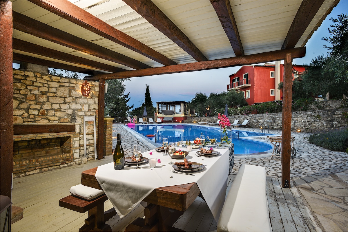 An outdoor dining area is set with a large wooden table featuring plates and glasses, ready for a meal. The pool sparkles in the background, surrounded by stone pathways and lounge chairs. Warm lighting highlights the covered space, and lush greenery adds a natural touch.
