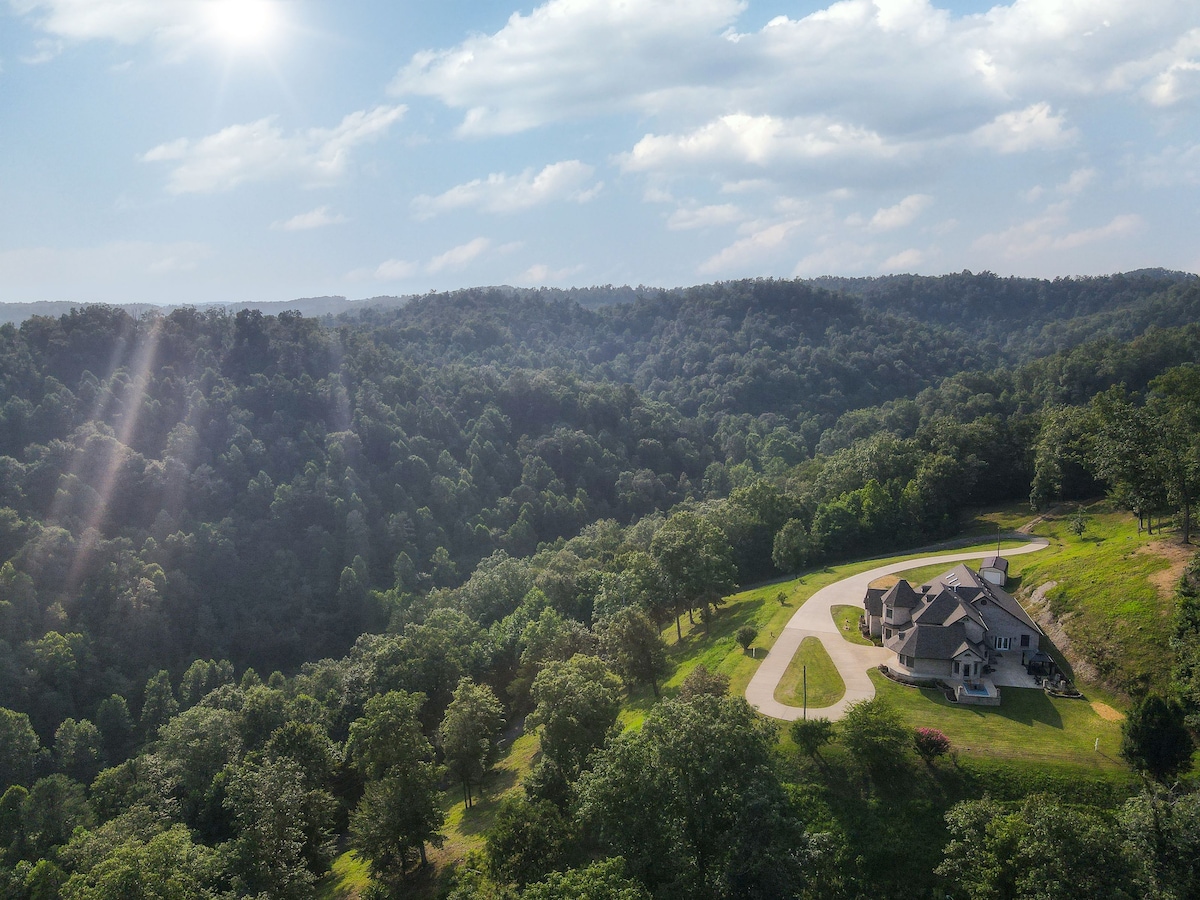 Aerial view captures the sprawling estate nestled among lush green hills, showcasing gentle slopes and dense tree coverage. The custom-built mansion is positioned prominently on a winding driveway, surrounded by scenic natural beauty under a bright, sunny sky.