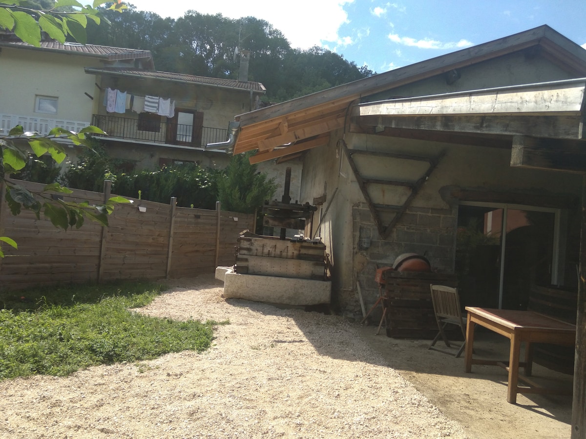 A gravel area surrounds a rustic outdoor space featuring a wooden table and chairs. A stone structure resembling a former wine press is positioned nearby, while laundry is visible hanging on a clothesline in the background. The setting is enclosed by a wooden fence and greenery.