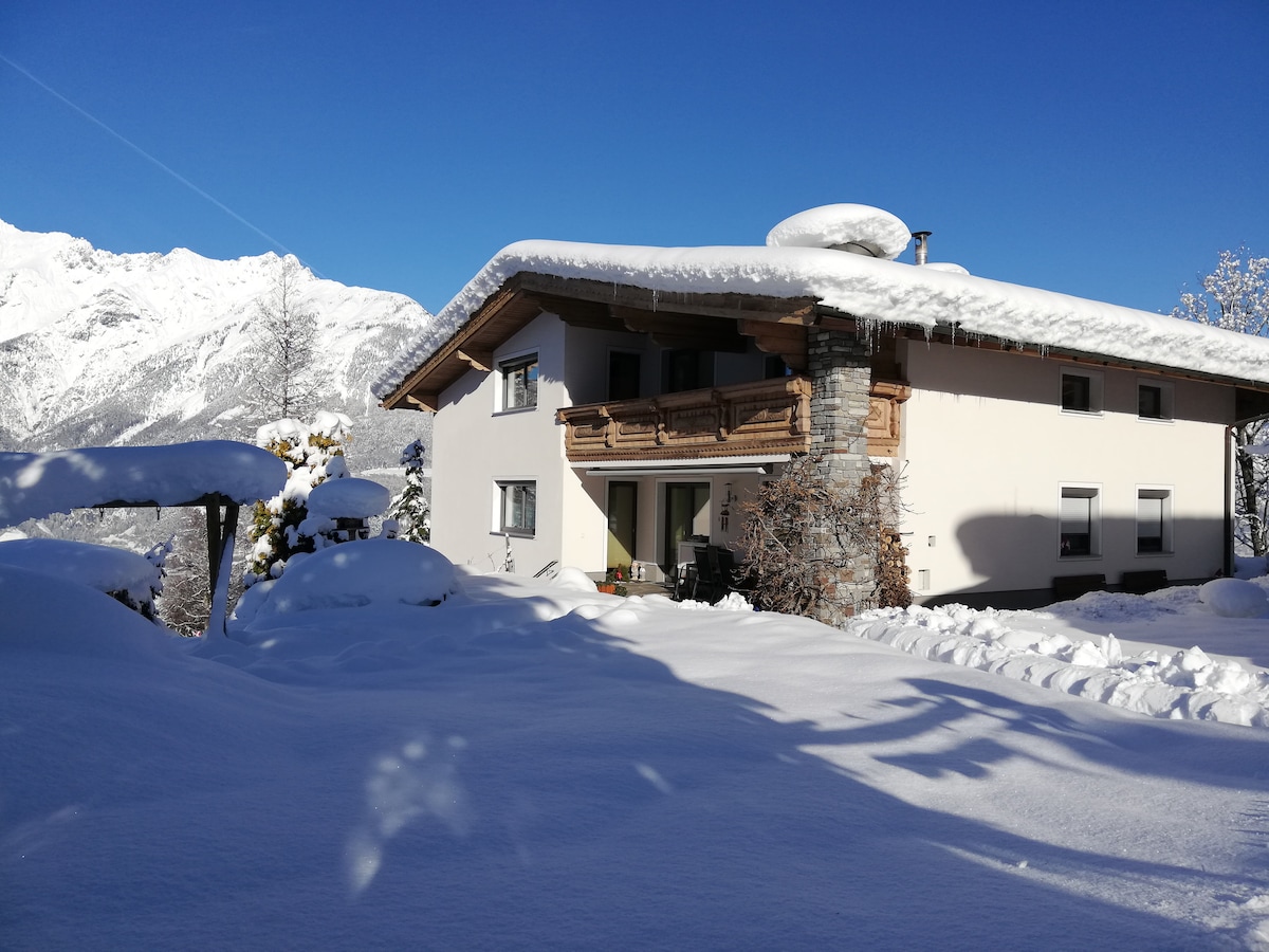 A spacious building is set against a backdrop of snow-covered mountains. Fresh snow blankets the ground, highlighting the architectural features, including a balcony adorned with wooden railings. Clear blue skies are visible, complementing the serene winter landscape.