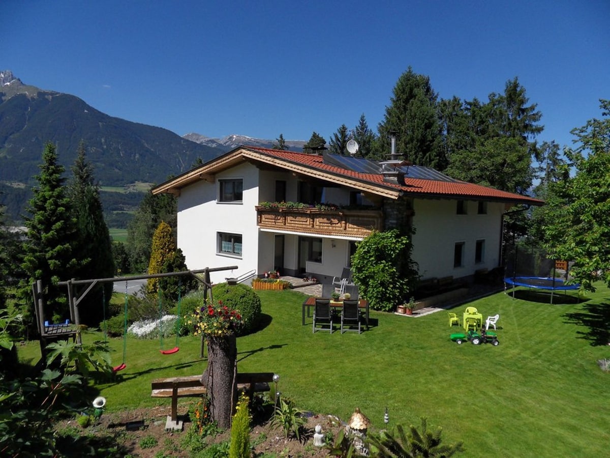 A two-story house is set in a lush green landscape with mountain views. The exterior features a balcony with garden furniture. A well-maintained lawn surrounds the house, while playful elements such as a trampoline and seating areas enhance the outdoor space.