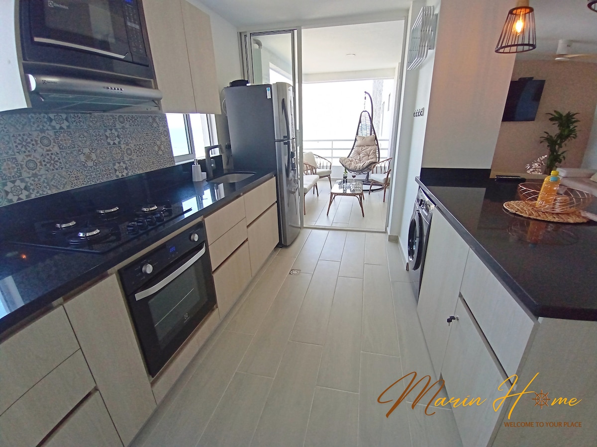 A modern kitchen is featured, equipped with a built-in oven, gas stove, and a refrigerator. Light wooden cabinetry contrasts with dark countertops. Large sliding glass doors lead to a balcony area, while a washing machine is visible on the side.