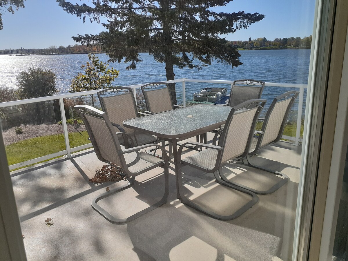 A spacious deck overlooks the shimmering lake, featuring a large dining table surrounded by eight lightweight chairs. Sunlight reflects off the water, enhancing the serene environment. The deck is bordered by a clear railing, providing unobstructed views of the tranquil surroundings.