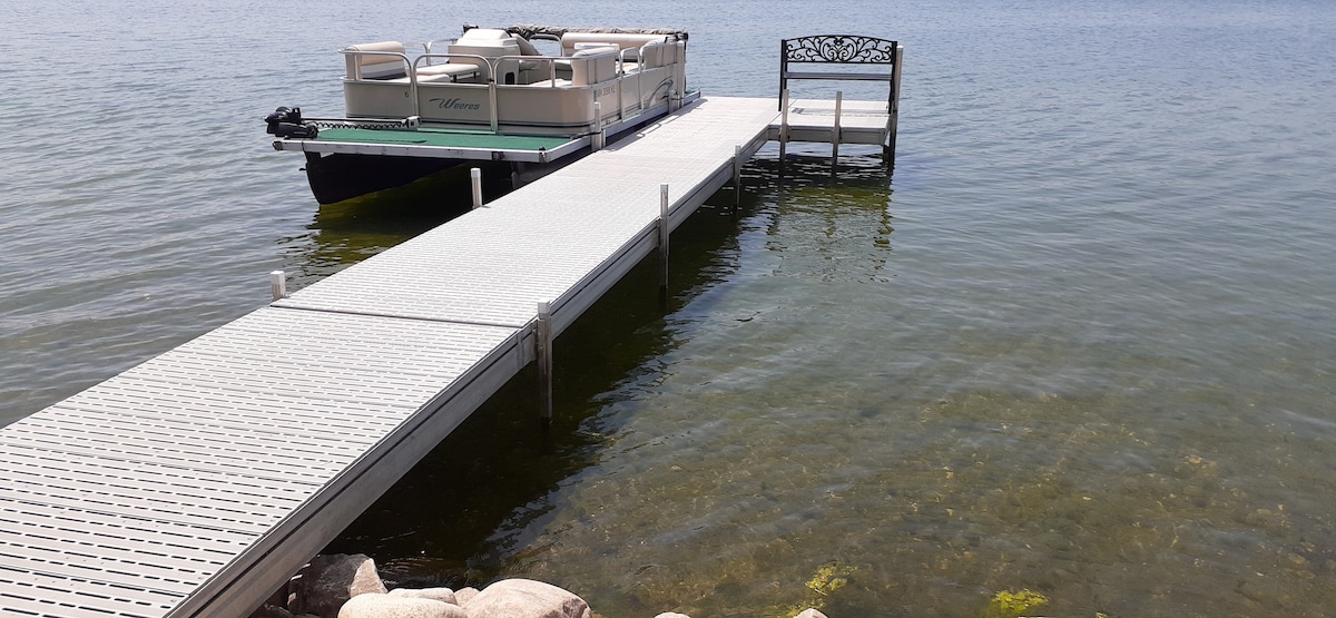 A floating pontoon boat is moored at the end of a white dock that extends over the calm, clear water. The dock features a bench at its edge, providing a resting place to enjoy the peaceful lake surroundings.