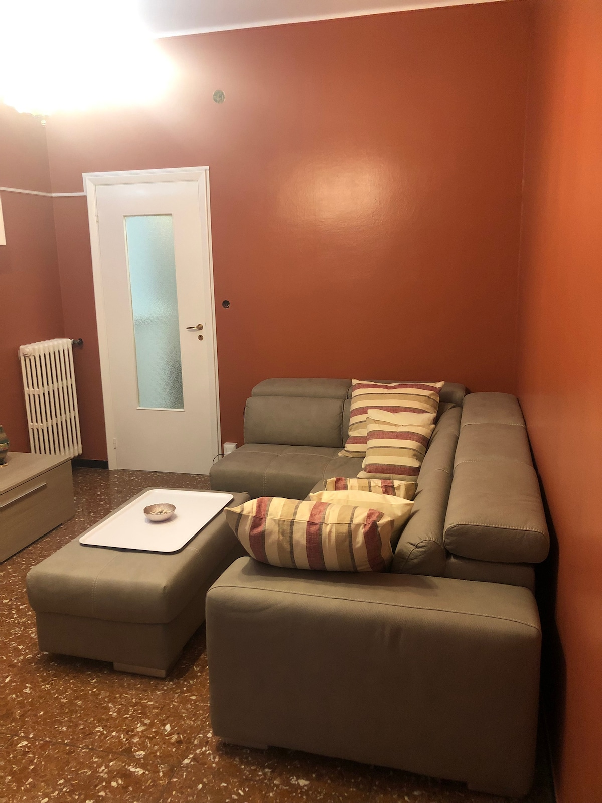 A sectional sofa is positioned in a cozy living area, featuring light-colored cushions with stripes. A small, rectangular table is placed in front of the sofa. An orange wall creates a warm backdrop, while a door and a radiator are visible in the space.