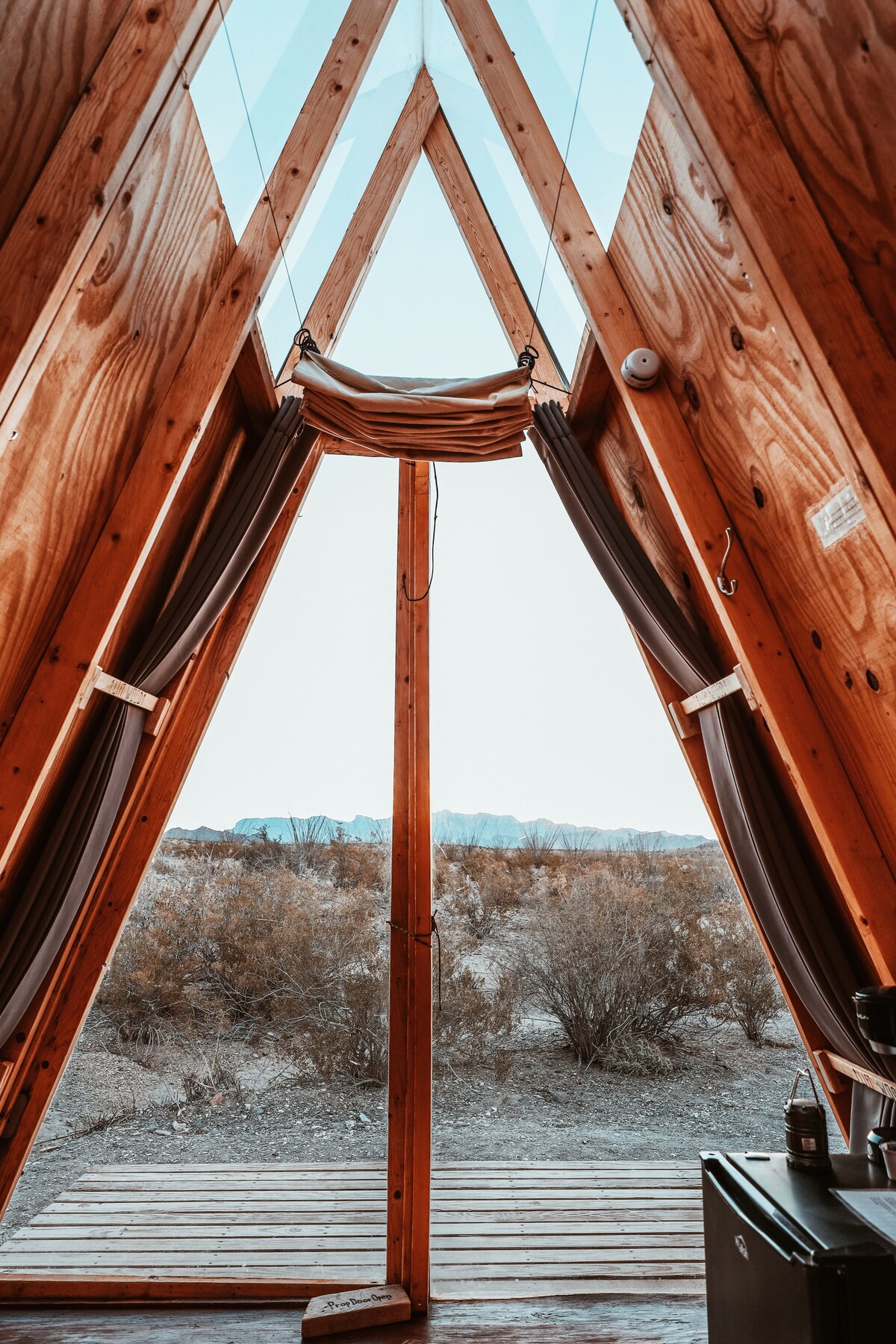 The image captures the interior view of an A-frame tent, featuring wooden walls and a ceiling of plexiglass that allows natural light to filter in. The entrance, framed by curtains, opens to a landscape with low shrubs and distant mountains, enhancing the outdoor feel.