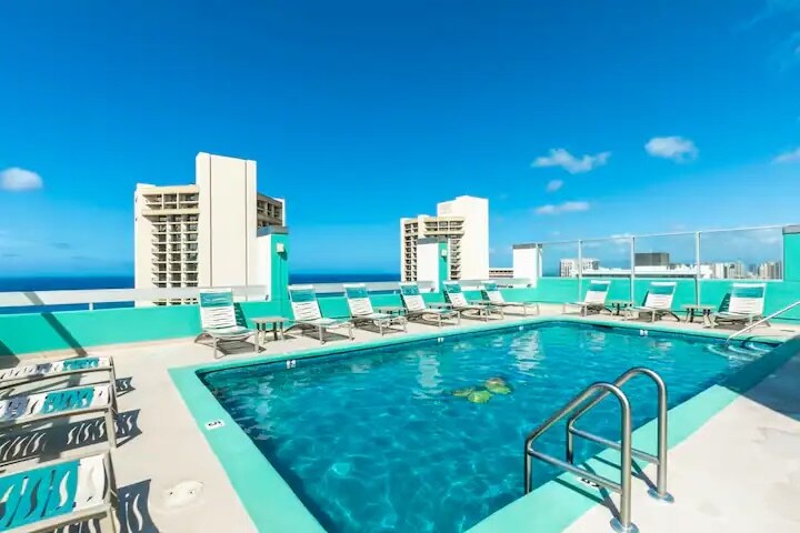 A rooftop pool area features clear blue water surrounded by lounge chairs. The space offers unobstructed views of the horizon and distant buildings against a bright blue sky.