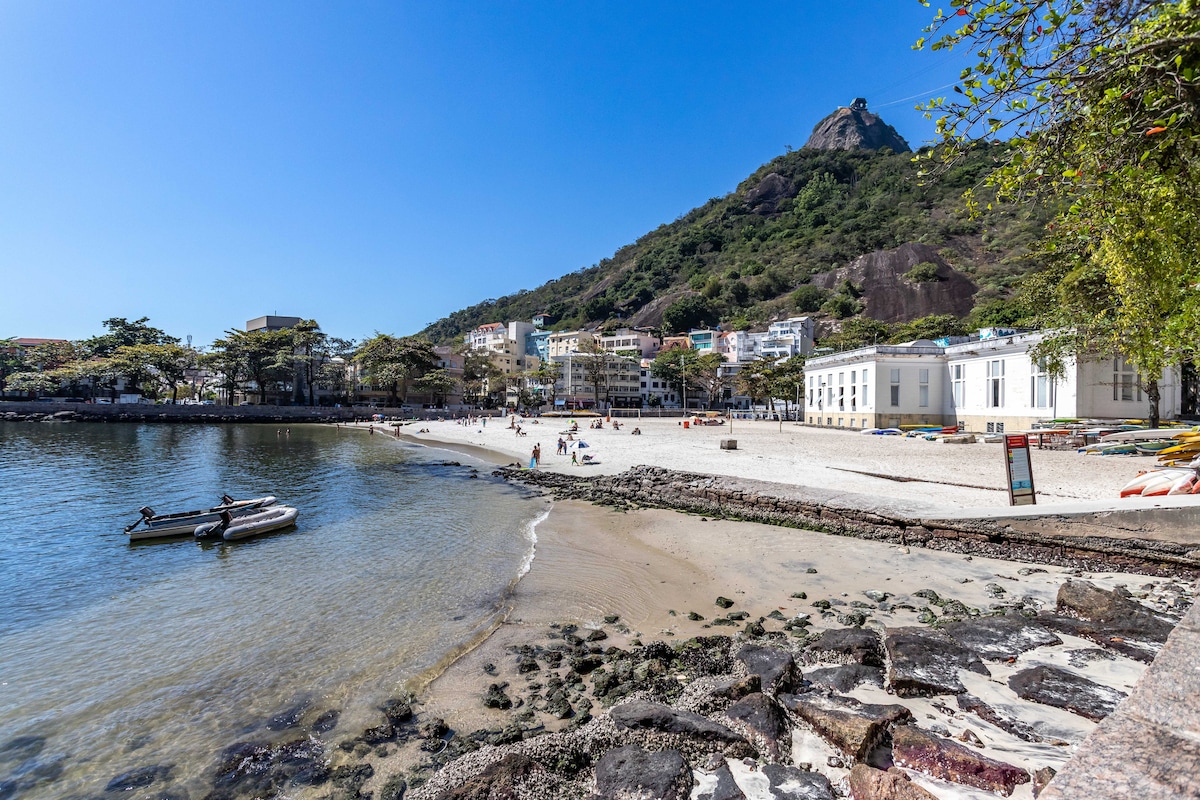 The image captures a sunny beach scene, featuring soft sand and clear waters. Small boats are anchored near the shore. The backdrop showcases a lush, green hill, while charming buildings line the beach, creating a serene atmosphere ideal for relaxation.