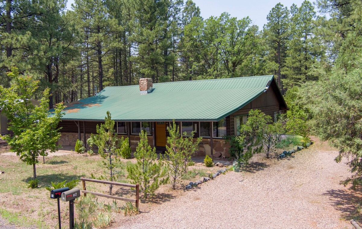 A cabin with a green metal roof is situated among tall pine trees. The exterior features stone accents and large windows, providing a glimpse of the surrounding nature. A gravel driveway leads to the entrance, with manicured landscaping visible in the foreground.