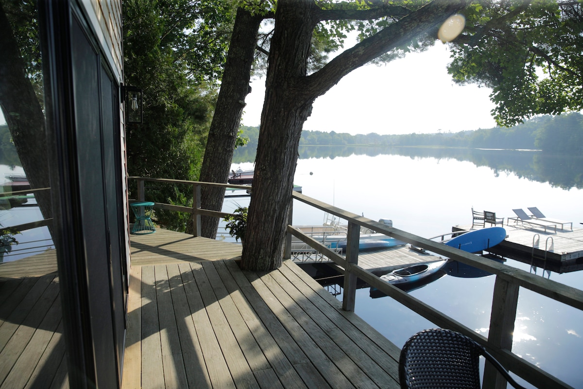 A wooden deck extends from the cottage, framed by a tree and overlooking Norton Pond. Reflections of the serene water and distant shoreline are visible. A large dock with chaise lounges and a canoe is nestled among the calm waters.