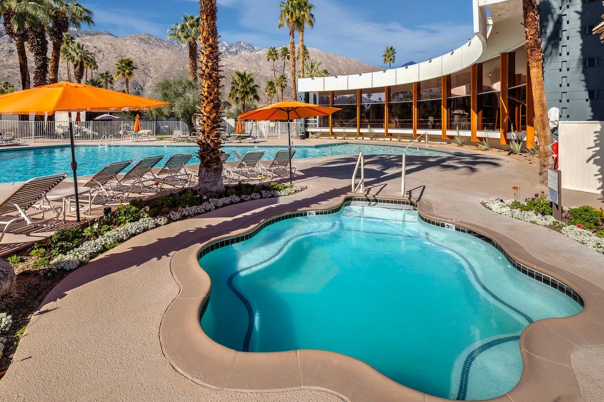 The pool area features a large swimming pool surrounded by lounge chairs under orange umbrellas. A hot tub is positioned nearby, with palm trees and mountains visible in the background. The smooth concrete surface complements the serene environment.