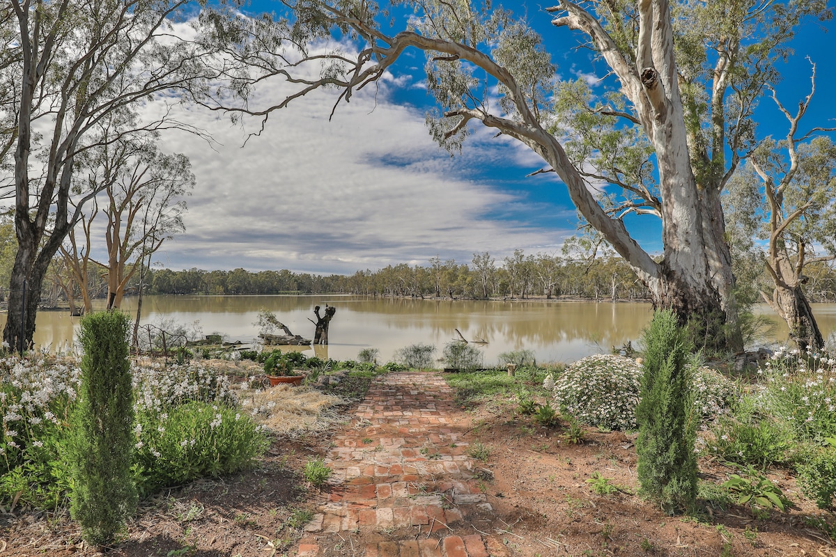 A scenic view of the Murray River is framed by tall trees and blooming wildflowers. A brick pathway leads from the homestead to the riverbank, inviting exploration and connection with nature. The sky features scattered clouds against a backdrop of blue, enhancing the tranquil ambiance.