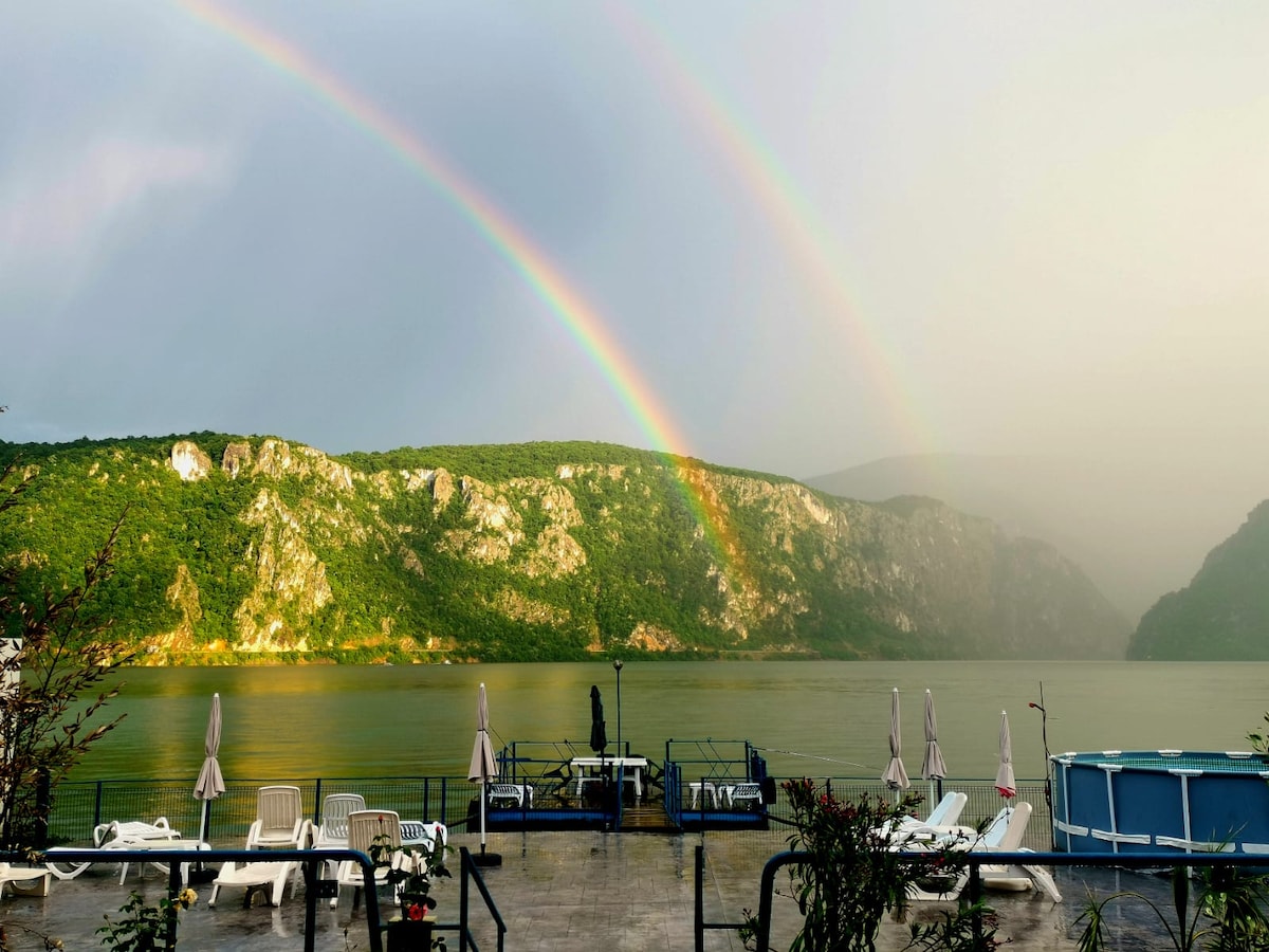 A serene landscape is depicted, featuring a calm body of water reflecting green hills. A double rainbow arcs over the scene, creating a vibrant display. Comfortable lounge chairs are positioned near the water's edge, inviting relaxation amidst the natural beauty.