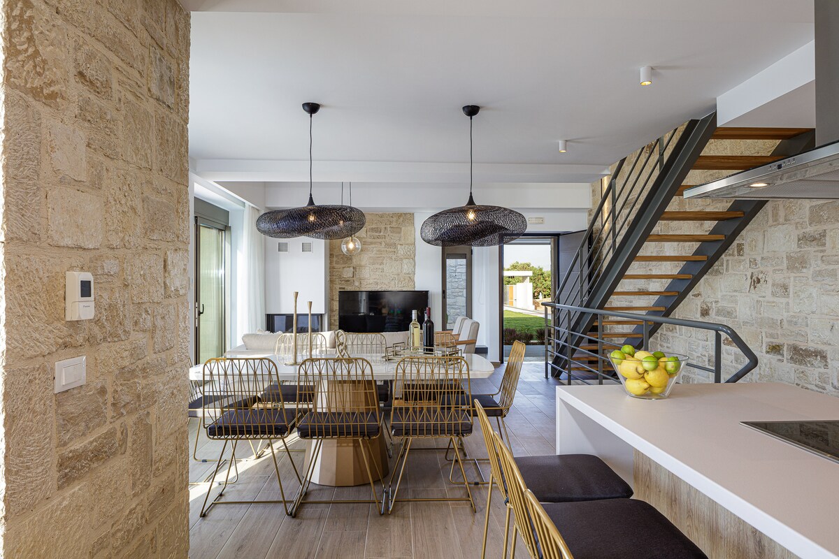 An open-plan living area features a contemporary dining table surrounded by gold wire chairs. Two pendant lights hang above the table, and a modern kitchen is visible with a bar counter. Large windows provide natural light and views of the outdoor space.