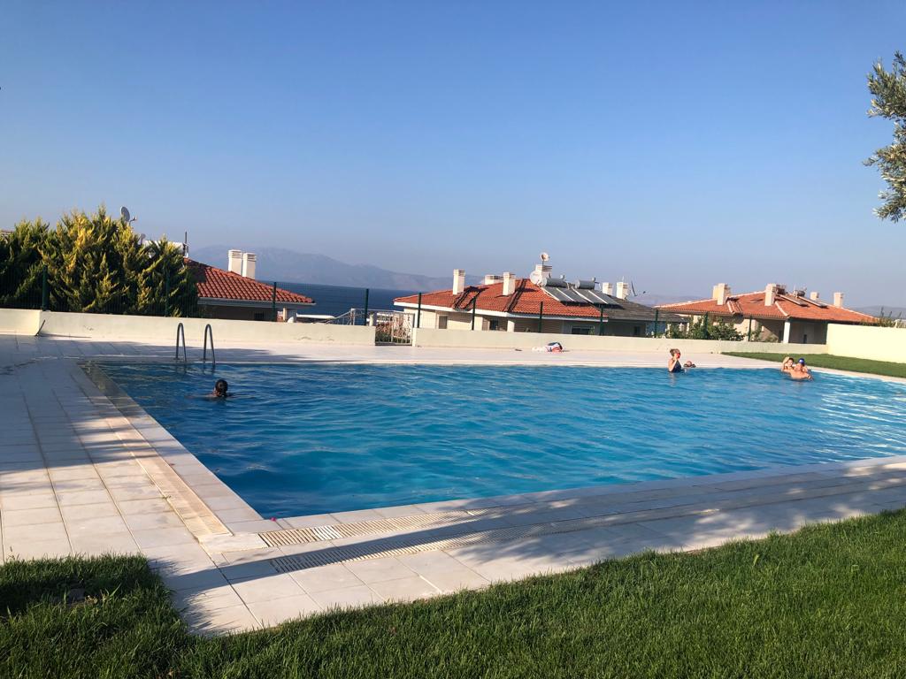 A tranquil swimming pool is set within a spacious outdoor area, surrounded by well-maintained grass. The clear blue water reflects the sky above, while rooftops and distant hills are visible in the background, creating a serene environment for relaxation.