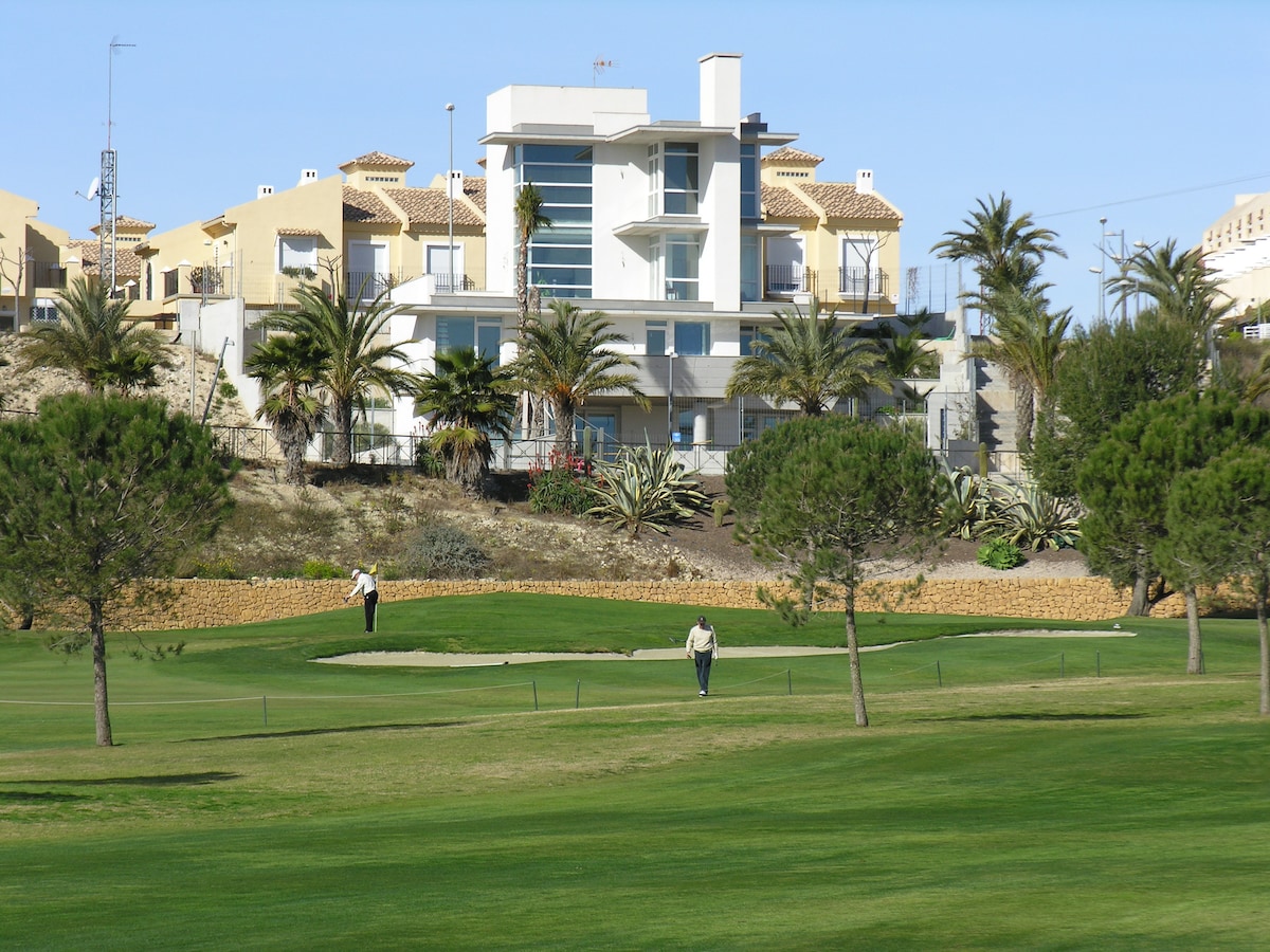 A landscaped golf course is visible with a manicured green expanse. Two individuals are seen on the fairway, with palm trees and lush plants lining the course. In the background, a modern residential building stands against a clear blue sky.