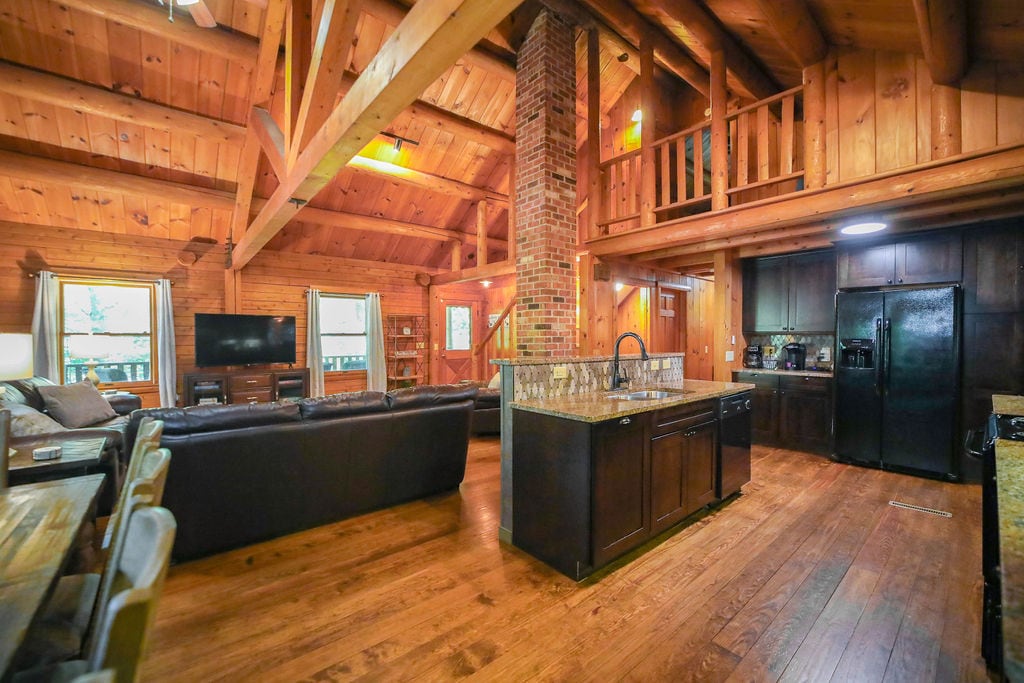 An open-concept living area showcases wooden beams and a vaulted ceiling. A cozy seating arrangement, featuring a large sectional sofa, is positioned near a mounted television. The kitchen, with dark cabinets and a stone backsplash, is easily accessible, fostering a welcoming atmosphere for gatherings.