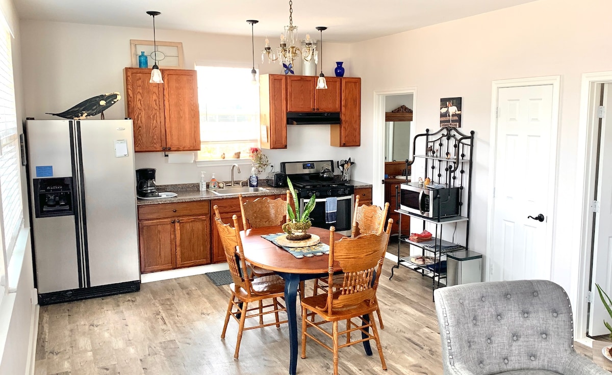 The kitchen area features wooden cabinetry and a countertop equipped with modern appliances. A round dining table is surrounded by four wooden chairs, and natural light filters through a window above the sink. Decorative items and plants add a homey touch.