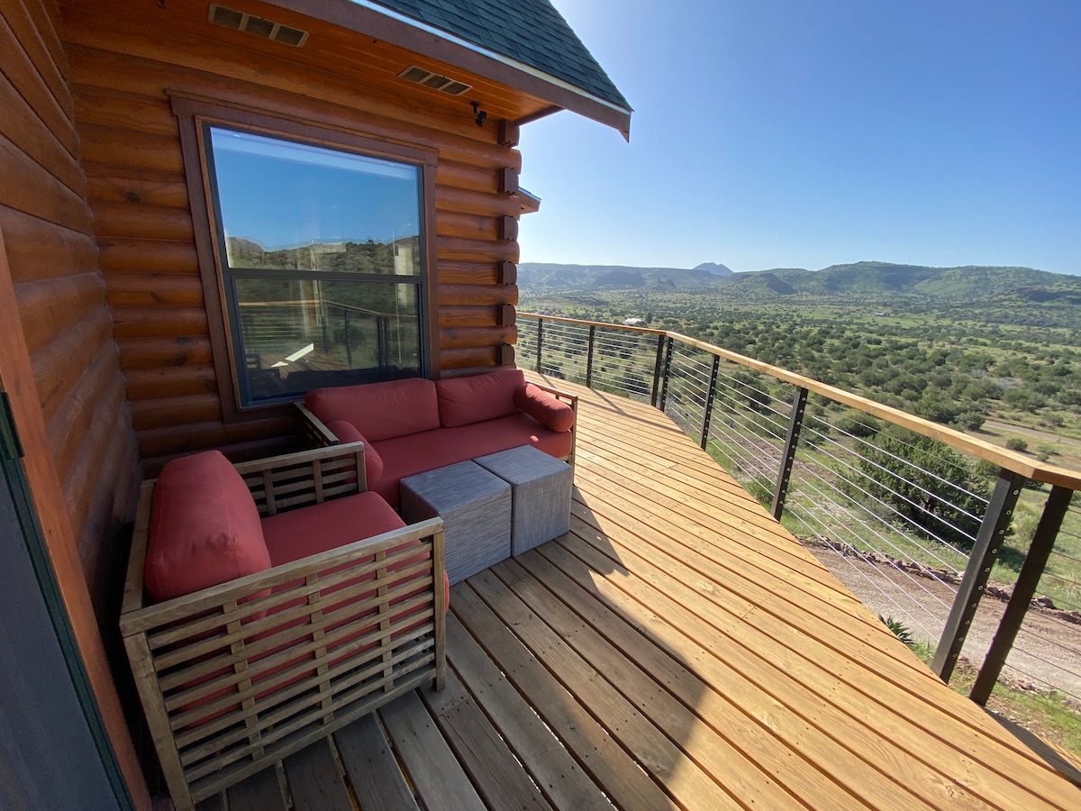 A wrap-around deck features comfortable red seating and a wooden coffee table, providing an ideal space for relaxation. Panoramic views of the surrounding hills and lush landscape are visible, complemented by clear blue skies above.