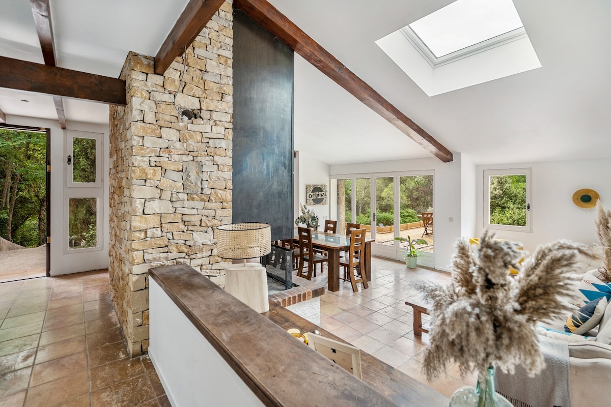 An open living area is highlighted by natural stone and wooden beams. Large windows invite natural light, with views of the surrounding greenery. A dining table for six is set against a backdrop of nature, while decorative elements add a touch of warmth.