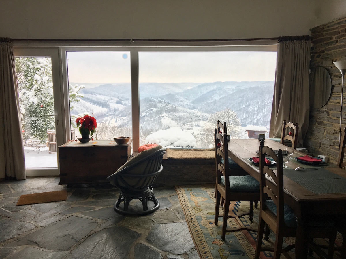 A spacious dining area with a wooden table is complemented by rustic chairs and a decorative bowl. A large window offers a panoramic view of a snowy landscape, framed by soft curtains, while a cozy seating area is visible nearby.
