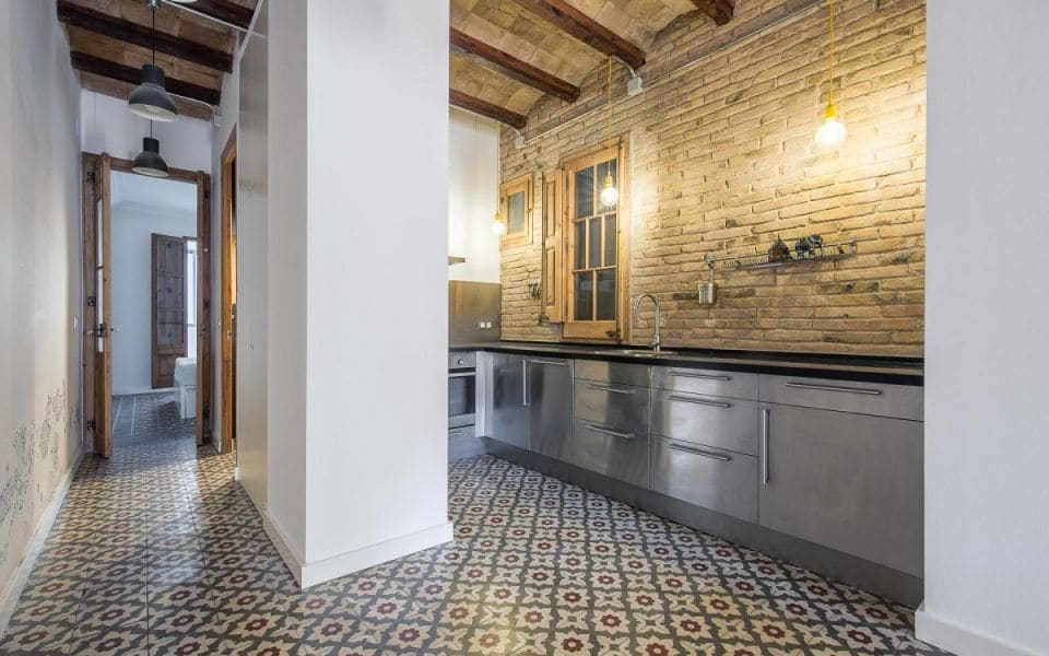 An open kitchen area showcases modern stainless-steel appliances against a backdrop of exposed brick walls. The floor features intricate hydraulic tiles, while pendant lights provide illumination. The space is complemented by large windows, bringing in natural light.
