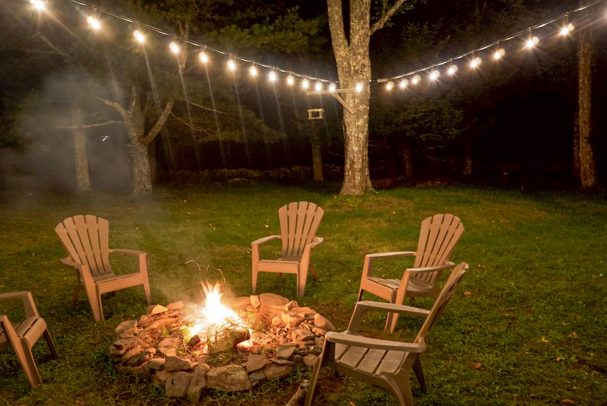 An outdoor fire pit surrounded by several Adirondack chairs is illuminated by string lights overhead. The grassy area features trees in the background, creating a serene atmosphere for gathering and relaxation during the evening.