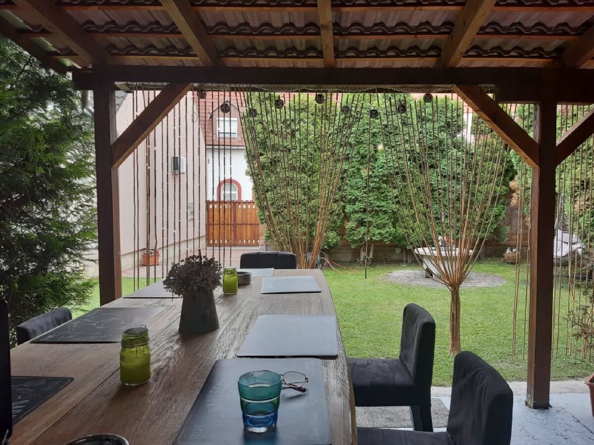 A dining area is situated under a wooden pergola, featuring a large table surrounded by dark chairs. Green plants line the yard, creating a serene atmosphere. Decorative elements, including a vase and glassware, are positioned on the table, highlighting the outdoor dining space.
