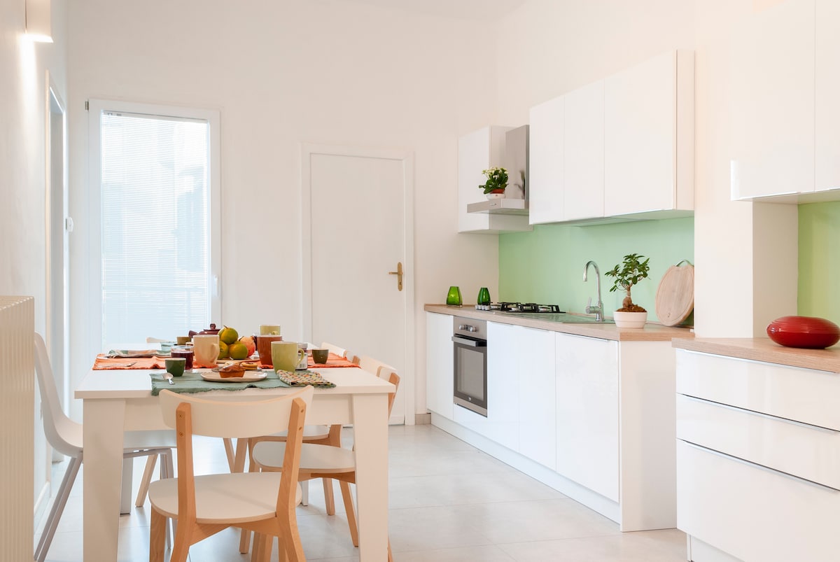 A modern kitchen is presented with white cabinetry and green accents. An inviting dining table is set for four, featuring plates and fruit. A stainless steel oven and a small potted plant are visible on the countertop, while natural light pours in through a nearby window.
