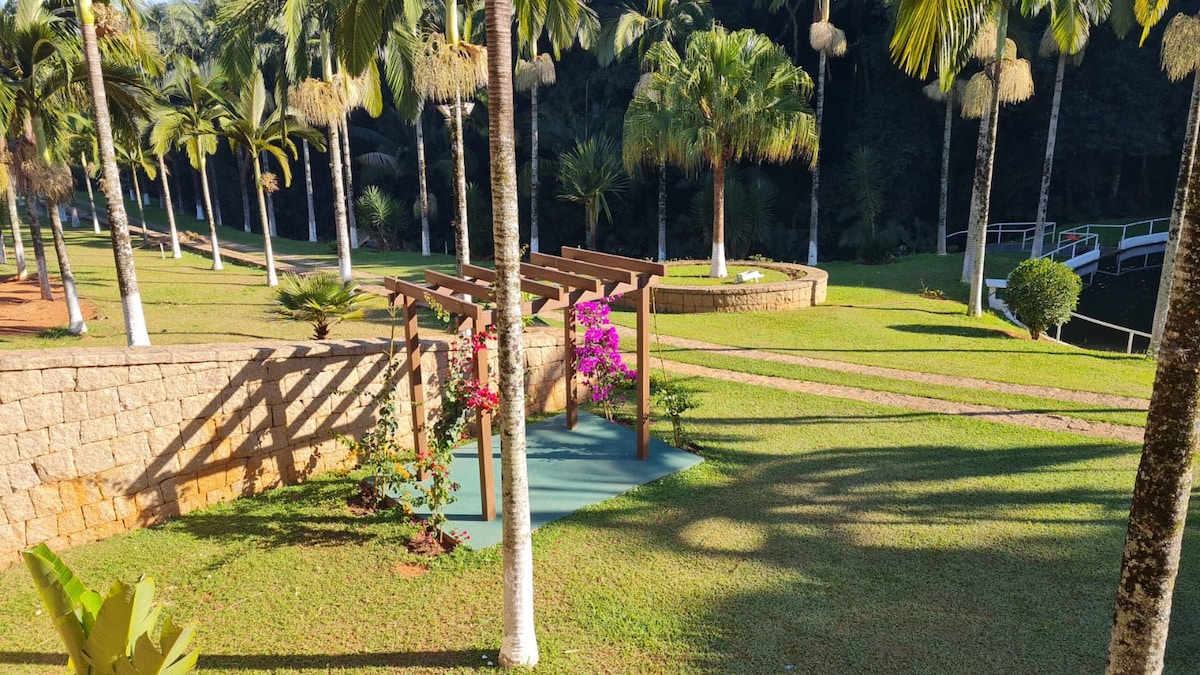 A landscaped garden area features neatly trimmed grass and palm trees. A wooden pergola, adorned with flowering vines in vibrant colors, offers a shaded spot. Pathways meander through the greenery, providing access to the surrounding natural beauty.
