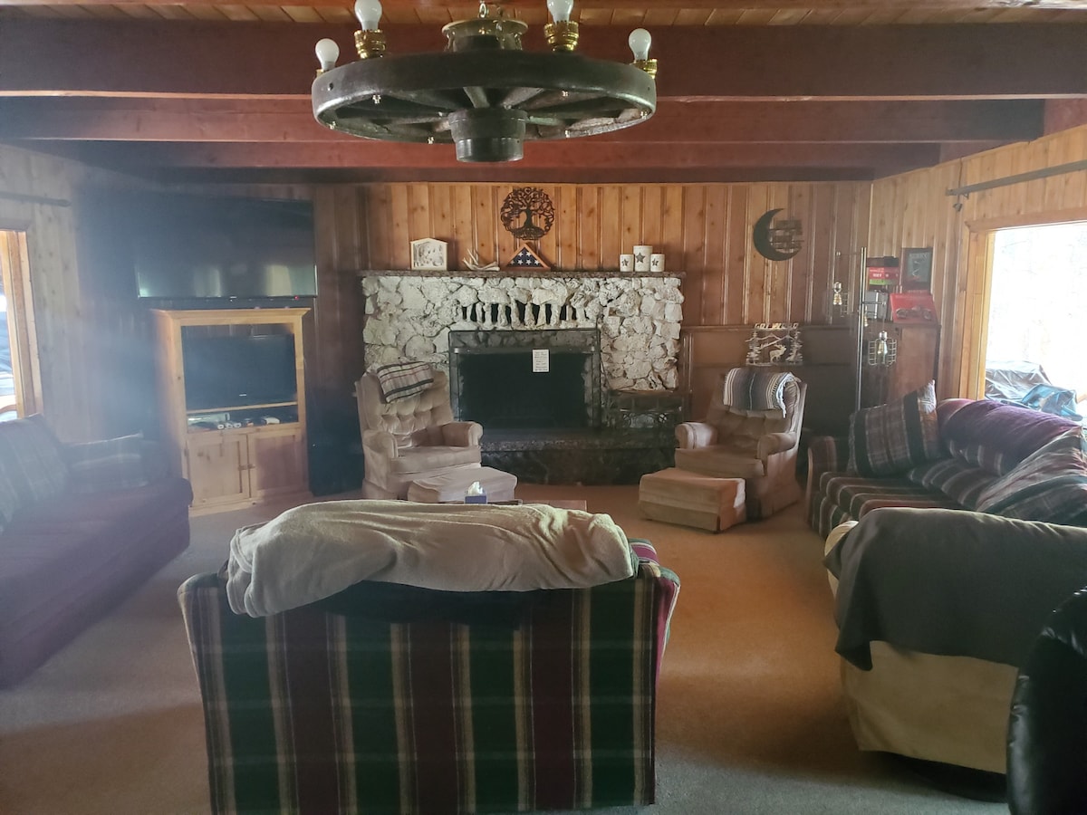A cozy living room is featured, showcasing wooden walls and a stone fireplace as a focal point. Comfortable seating options, including plaid-patterned sofas and armchairs, are arranged for relaxation. Large windows allow natural light to fill the space, offering views of the surrounding woods.